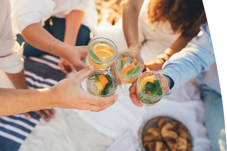 Group of happy friends having picnic, drinking lemonade and eating fruits, sitting near sea at sunset